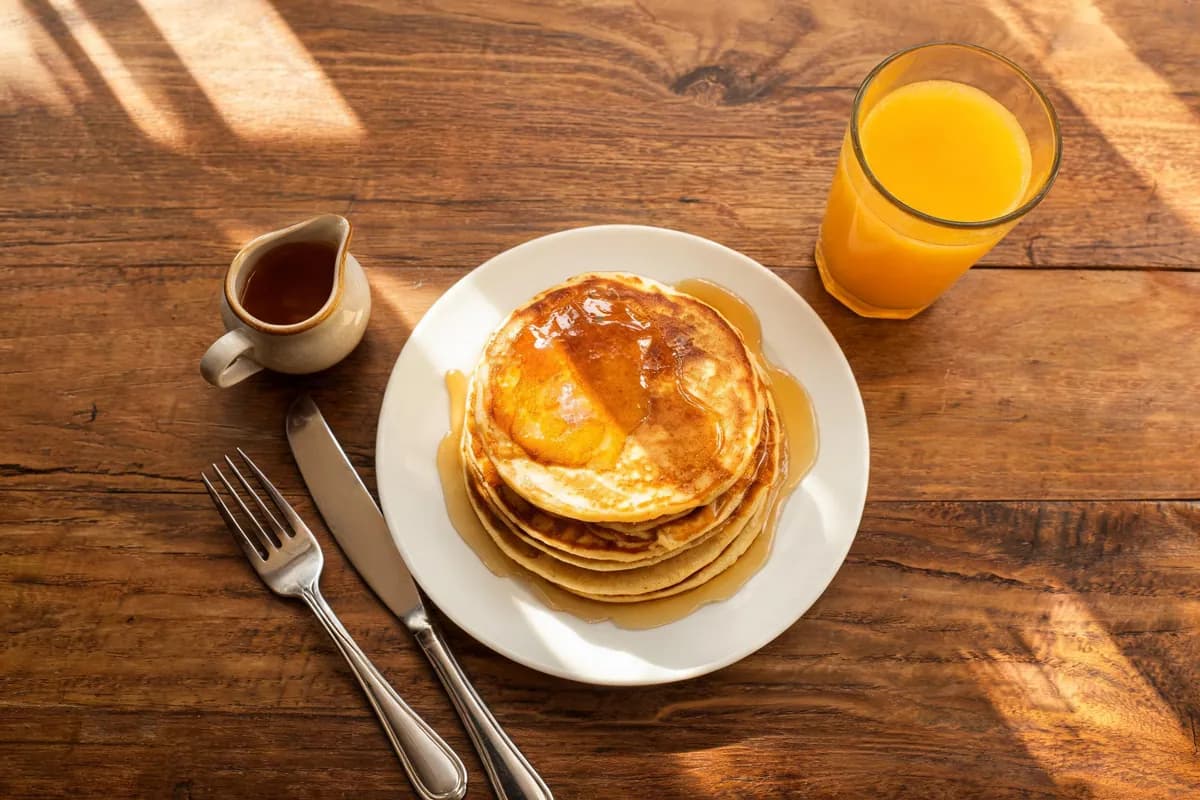 Input: a plate of pancakes with a glass of orange juice and a jug of syrup on a wooden table.